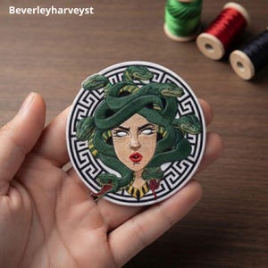 May include: A circular embroidered patch featuring a central figure with white eyes and red lips, surrounded by green snakes. The design is framed by a black and white Greek key border. The patch is held against a wooden background with thread spools.