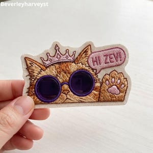 Op de afbeelding: Een geborduurde patch met een oranje kat die een lichtroze kroon en een donkerpaarse ronde zonnebril draagt. De kat heeft een opgeheven poot en een tekstballon met de tekst HI ZEV!. De patch is op een crèmekleurige stoffen ondergrond.