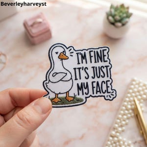 May include: An embroidered patch featuring a white duck with an orange beak and feet, standing with its wings crossed. The text reads, I'M FINE IT'S JUST MY FACE, in black lettering. The patch has a dark blue border and a small patch of green grass.