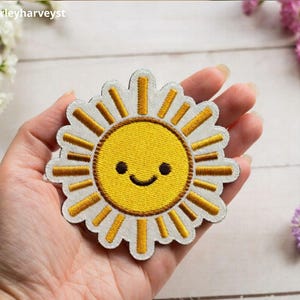 May include: An embroidered patch shaped like a sun with a bright yellow center, a smiling face with two black eyes and a curved mouth, and white scalloped edges with radiating yellow sunbeams extending outward from the central circle.