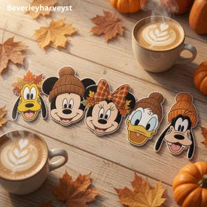 May include: Five embroidered patches featuring cartoon character faces wearing autumn-themed hats and accessories, including Pluto, Mickey, Minnie, Donald, and Goofy, arranged on a wooden surface with coffee mugs and orange pumpkins.