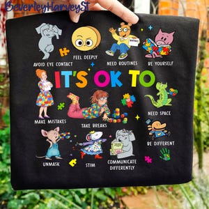May include: Black t-shirt with colorful illustrations and the text "IT'S OK TO." The design includes cartoon characters and phrases like "Avoid Eye Contact," "Feel Deeply," and "Make Mistakes." The shirt promotes inclusivity and self-acceptance.