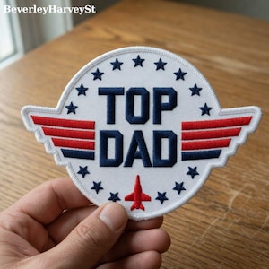 May include: A white patch with the text "TOP DAD" in navy blue, surrounded by stars. Red and navy blue wings are on either side, and a red airplane is at the bottom. The patch is held by a hand.