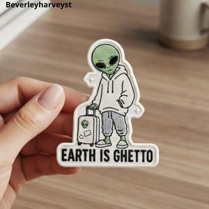 May include: An embroidered patch featuring a green alien wearing a white hoodie and gray sweatpants, standing next to a small rolling suitcase with an alien sticker. The text "EARTH IS GHETTO" is printed in black capital letters at the bottom.
