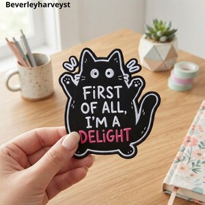 以下が含まれることがあります： 白い輪郭と大きな目を持つ、猫の形をした黒い刺繍ワッペン。テキストは「FIRST OF ALL, I'M A DELIGHT」と書かれており、「DELIGHT」という単語はピンク色の刺繍で施されています。猫は前足を上げ、尻尾を曲げた平らなデザインです。