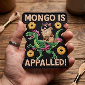 Pode incluir: Patch preto com o texto "MONGO IS APPALLED!" apresentando um dinossauro verde com detalhes em rosa e um gato usando óculos de sol e uma coroa. O patch também inclui gráficos de donuts.
