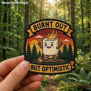 May include: An embroidered patch featuring a cartoon marshmallow with a flame on its head, standing in a forest scene with a sunset. The text BURNT OUT is at the top and BUT OPTIMISTIC is at the bottom in bold black letters on orange banners.