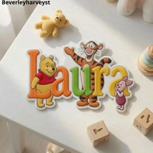 Puede incluir: Un letrero decorativo con el nombre Laura en letras coloridas y en relieve, con Winnie the Pooh, Tigger y Piglet. Las letras son de color naranja, verde y amarillo, con los personajes integrados en el diseño. El letrero está sobre una superficie blanca.
