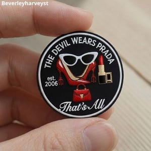 May include: A circular black embroidered patch featuring white text that reads THE DEVIL WEARS PRADA, est. 2006, and That's All. The design includes white cat-eye sunglasses, red high-heeled shoes, a red handbag, and a red lipstick tube.