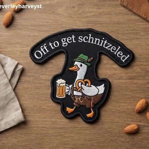 May include: A black embroidered patch featuring a white goose wearing a green alpine hat and brown lederhosen. The goose holds a beer mug and a pretzel. The text "Off to get schnitzeled" is arched in white lettering above the goose illustration.
