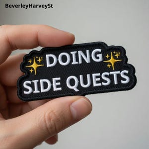 Puede incluir: Un parche bordado negro con las palabras "DOING SIDE QUESTS" en blanco, texto estilizado. El parche tiene un borde negro y detalles de estrellas amarillas. El parche se sostiene en una mano.