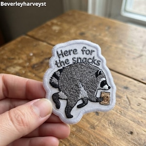 May include: A white embroidered patch featuring a gray and black raccoon holding a small tan snack bag. The text "Here for the snacks" is stitched in black thread above the raccoon. The patch has a white border and a die-cut shape.