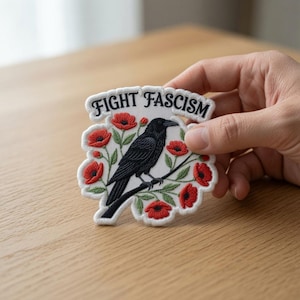 May include: An embroidered patch with a white background featuring a black crow perched on a branch surrounded by red poppy flowers and green leaves. The text "FIGHT FASCISM" is embroidered in black capital letters along the top edge of the patch.