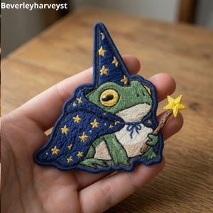 May include: An embroidered patch featuring a green frog wearing a dark blue wizard hat and cape decorated with yellow stars. The frog holds a brown wand topped with a yellow star. The text Beverleyharveyst is visible in the top left corner.