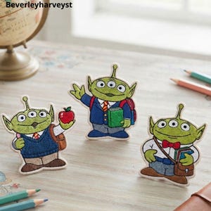 Puede incluir: Tres parches bordados que muestran alienígenas verdes de tres ojos vestidos como estudiantes. Uno sostiene una manzana roja, otro lleva un libro verde y una mochila roja, y el tercero usa una pajarita roja con un bolso de cuero marrón.