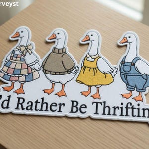 May include: A white patch featuring four geese dressed in various outfits. The geese are wearing a dress, sweater, yellow dress, and overalls. The text "I'd Rather Be Thrifting" is below the geese.