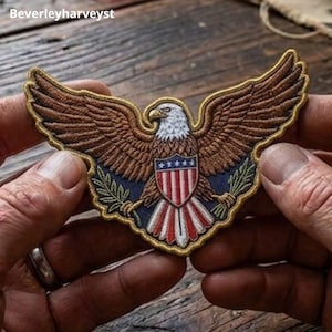 May include: An embroidered patch featuring a bald eagle with outstretched brown wings, a white head, and a shield on its chest displaying red and white vertical stripes with a blue top section containing white stars, bordered by gold thread.
