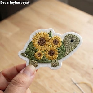 Puede incluir: Un parche bordado con forma de tortuga cuyo caparazón presenta tres girasoles amarillos con centros marrones y hojas verdes. La tortuga tiene un cuerpo texturizado de color verde claro y un solo ojo negro. El texto Beverleyharveyst es visible.