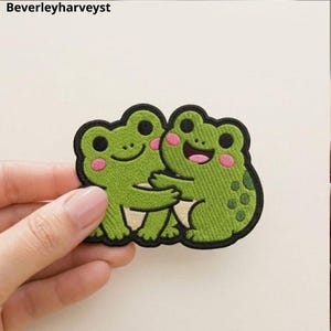 Puede incluir: Un parche bordado que muestra a dos ranas verdes abrazándose. Las ranas tienen ojos negros, mejillas rosadas y vientres claros. El parche tiene un borde negro grueso y el texto Beverleyharveyst es visible en la esquina superior izquierda.
