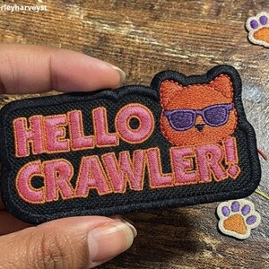 May include: A rectangular embroidered patch with a black background featuring the pink text HELLO CRAWLER! and an orange cat head wearing purple sunglasses. Two small paw print patches with orange centres and purple toes are visible nearby.