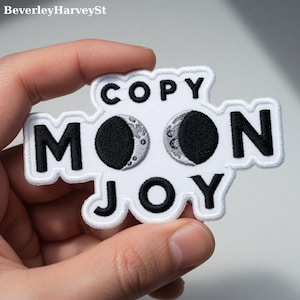 Puede incluir: Un parche bordado blanco con las palabras "COPY MOON JOY" en letras negras. La palabra "MOON" presenta dos diseños de luna creciente en blanco y negro. El parche tiene un borde blanco.