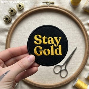 May include: A circular black embroidered patch featuring the yellow text Stay Gold in a bold serif font. The patch is held against a background containing a wooden embroidery hoop, metal bobbins, silver scissors, and yellow sewing thread.