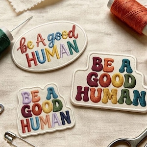 May include: Three embroidered fabric patches with off-white backgrounds, each featuring the text "be A good HUMAN" in multi-coloured, rounded, puffy-style lettering. The patches vary in shape, including one oval and two irregular shapes.