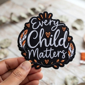 May include: A black embroidered patch featuring the white text Every Child Matters in a script font. The design includes orange and white feather motifs on the sides, small orange heart outlines, and orange leaf accents at the bottom edge.