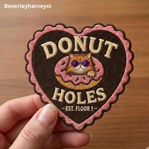 Peut inclure: Un écusson brodé en forme de cœur avec une bordure rose festonnée et un fond marron foncé. Il représente un chat roux portant des lunettes de soleil violettes à travers un donut glacé rose. Le texte indique DONUT HOLES EST. FLOOR 1.