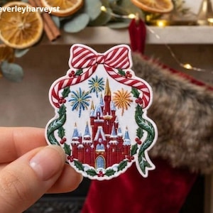 May include: A white embroidered patch featuring a red castle with blue accents, surrounded by a green holly wreath with red berries and topped with a red and white striped bow. The design includes colourful fireworks in the background.