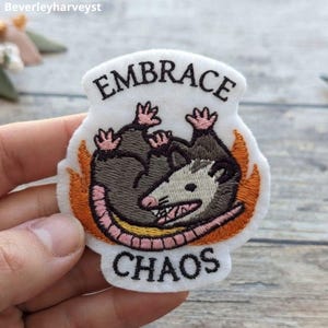 May include: An embroidered patch on a white background featuring a grey and white opossum curled up inside orange flames. The text EMBRACE is stitched in black above the opossum, and the text CHAOS is stitched in black below the opossum.