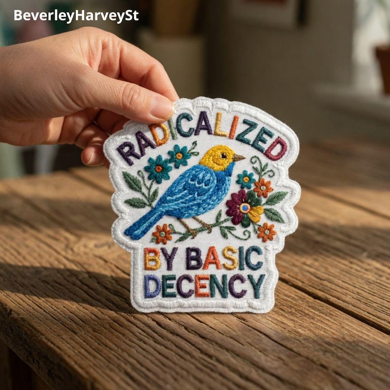 Op de afbeelding: Geborduurde patch met een blauwe vogel met gele kop, omringd door kleurrijke bloemen en de tekst "RADICALIZED BY BASIC DECENCY". De patch heeft een witte geschulpte rand.