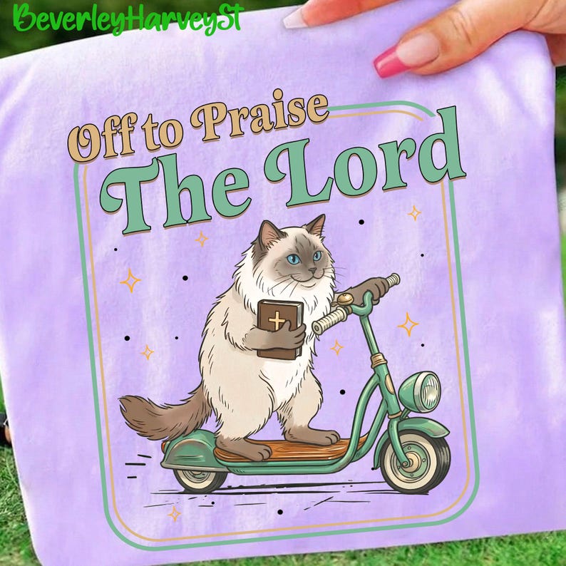 May include: A light purple t-shirt featuring a cartoon cat riding a green scooter and holding a book with a cross. The text "Off to Praise The Lord" is above the cat. The design is framed with a light green and orange border.