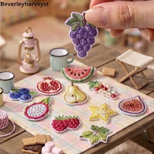 May include: A collection of small, embroidered fruit patches featuring a purple grape cluster, watermelon slice, pomegranate, pear, dragon fruit, raspberries, starfruit, fig, and gooseberries arranged on a plaid fabric surface.