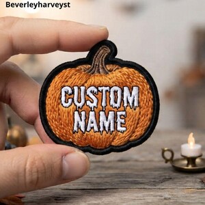 Könnte beinhalten: Ein gestickter kürbisförmiger Aufnäher mit schwarzem Rand. Der Kürbis ist orange mit einem braunen Stiel und zeigt den weißen Text "CUSTOM NAME" in einer tropfenden, gruseligen Schriftart in der Mitte der Aufnäheroberfläche.