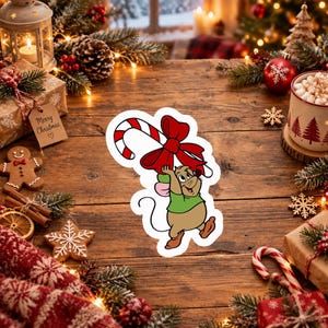 Op de afbeelding: Een gestanste sticker met een getekende muis met lichtbruine vacht, gekleed in een groen shirt en bruine schoenen, die een grote rood-wit gestreepte zuurstok met een opvallende rode strik boven het hoofd houdt op een witte achtergrond.
