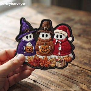 May include: An embroidered patch featuring three white ghost figures. The left ghost wears a purple witch hat and jumper, holding a pumpkin mug. The centre ghost wears a brown hat and jumper, holding a pumpkin. The right ghost wears a red Santa hat and jumper, holding a candy cane. Autumn leaves and gifts are at the base.