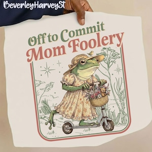 May include: A whimsical illustration features a frog riding a scooter, wearing a dress and hat, with a basket of flowers. The text reads "Off to Commit Mom Foolery" in green and red. The artwork has a vintage, storybook style.