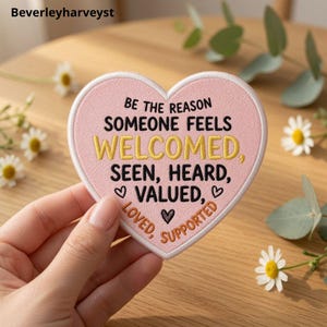 May include: A heart-shaped patch with a light pink background and white border. The text reads: BE THE REASON SOMEONE FEELS WELCOMED, SEEN, HEARD, VALUED, LOVED, SUPPORTED. The words are embroidered in black, yellow, and orange with small heart icons.