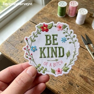 May include: A circular white embroidered patch with a scalloped edge featuring the green text BE KIND and the pink text OF A BITCH. The design is surrounded by an embroidered wreath of small red, pink, and blue flowers with green leaves and stems.