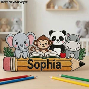May include: A decorative wooden cutout featuring an embroidered-style design of a gray elephant, a brown monkey, a black and white panda, and a gray hippo sitting on a large yellow pencil. The name Sophia is printed in black on the pencil.