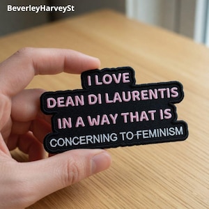 Pode incluir: Um patch bordado preto com texto rosa que diz "I LOVE DEAN DI LAURENTIS IN A WAY THAT IS CONCERNING TO FEMINISM." O patch tem uma borda preta e é segurado contra uma superfície de madeira clara.