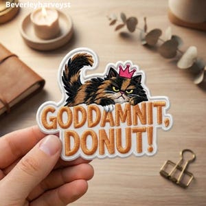 May include: An embroidered patch featuring a grumpy calico cat wearing a small pink crown. The cat is positioned above the text "GODDAMNIT, DONUT!" stitched in bold orange letters on a white background with a white border outline.