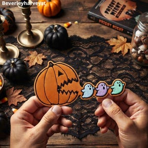 Puede incluir: Un recorte plano con forma de calabaza naranja y una boca abierta dentada. De la boca salen tres pequeños recortes con forma de fantasma en azul claro, morado claro y verde claro. La calabaza presenta detalles de contorno en negro.