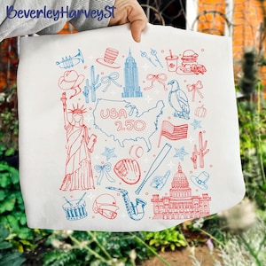 Könnte beinhalten: Weiße Tragetasche mit roten und blauen Illustrationen amerikanischer Symbole, darunter die Freiheitsstatue, das Empire State Building und eine Karte der USA mit dem Text "USA 250".
