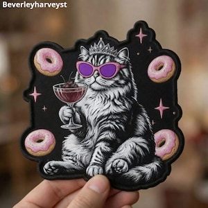 May include: A black embroidered patch featuring a white and grey tabby cat wearing a silver crown and purple cat-eye sunglasses. The cat holds a cocktail glass with cherries, surrounded by four pink frosted donuts and small pink star accents.