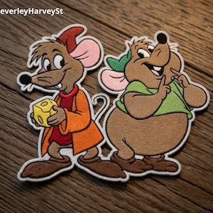 May include: Embroidered patches featuring two cartoon mice. One mouse is brown with an orange coat and red shirt, holding a yellow cheese. The other mouse is brown with a green shirt and a green hat. Both have pink ears and are smiling.