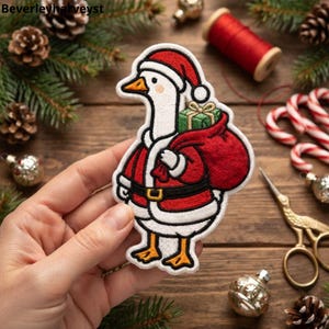 May include: An embroidered patch featuring a white goose wearing a red Santa hat and a red Santa coat with a black belt. The goose carries a red sack over its shoulder containing a green gift box with a gold ribbon. The patch has a white border.