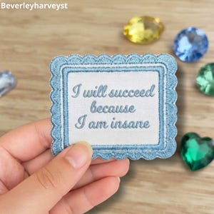 Può includere: Una toppa ricamata rettangolare con bordo smerlato in azzurro e bianco. Al centro è presente il testo 'I will succeed because I am insane' ricamato con filo blu. La toppa ha uno sfondo in tessuto bianco e un bordo di colore blu.