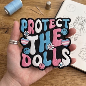 May include: Embroidered patch with the text "PROTECT THE DOLLS" in white, pink, and blue. The patch features smiley faces, flowers, and a transgender flag heart on a black background. A spool of blue thread, needles, and a thimble are in the background.